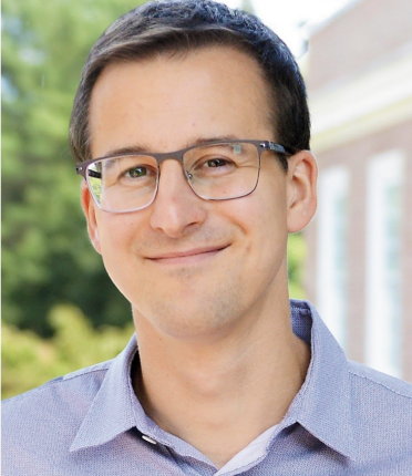 white man wearing glasses and blue collared shirt with dark hair smiling without showing teeth